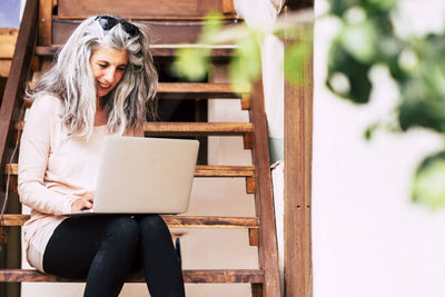 Smiling mature woman sitting on staircase while using laptop outdoors