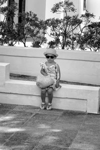 Full length of cute girl wearing hat and holding basket while sitting on retailing wall