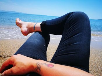 Low section of mature woman relaxing at beach against blue sky during sunny day