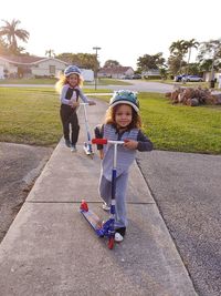 Portrait of smiling girls riding push scooters on footpath 