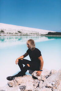 Man sitting on rock by lake against sky