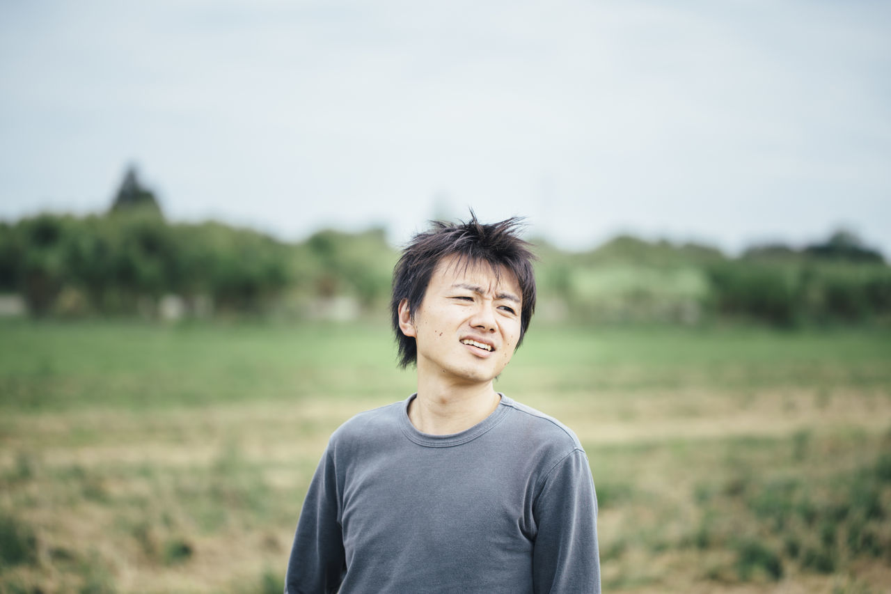 one person, smiling, portrait, emotion, nature, happiness, landscape, environment, rural scene, sky, waist up, casual clothing, field, focus on foreground, black hair, portrait photography, land, copy space, adult, outdoors, rural area, child, day, plant, men, front view, human face, standing, grass, person, young adult, childhood, tree, lifestyles