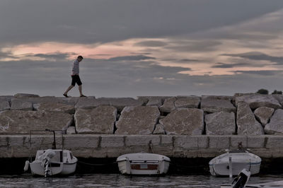 Side view of man walking on retaining wall by sea against cloudy sky during sunset