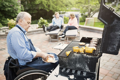 Disabled man barbecuing while family sitting in background at yard