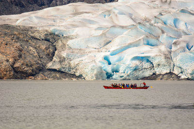 People sitting in rowboat on sea by glaciers