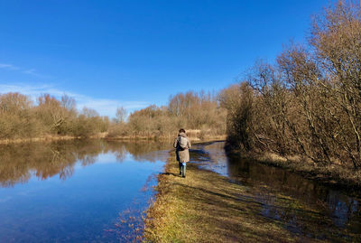 Rear view of a woman walking over a small dike between water and trees under a clear sky