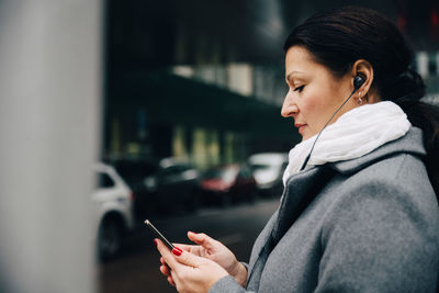 Side view of businesswoman listening music using mobile phone while standing on road in city