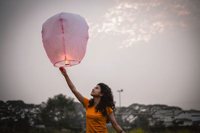 Woman holding umbrella with balloons against sky during sunset