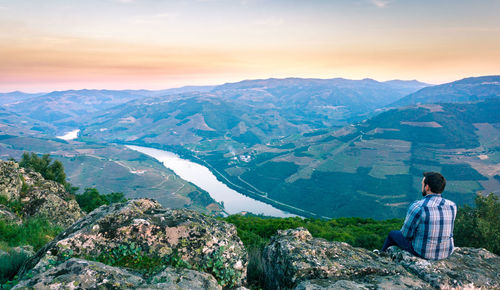 Rear view of man sitting on mountain against sky during sunset