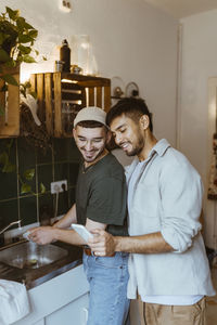 Smiling man washing dishes while boyfriend sharing smart phone in kitchen at home