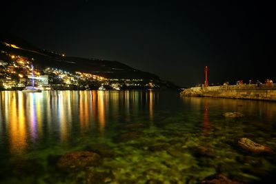 Illuminated buildings by sea against sky at night