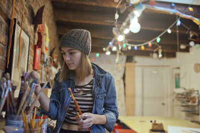 Young woman holding paintbrushes while standing at home