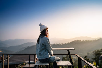 Woman sitting on railing against mountain during sunset