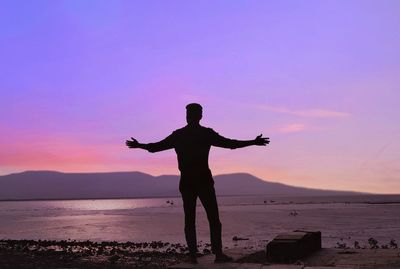 Rear view of silhouette man standing at beach against sky during sunset