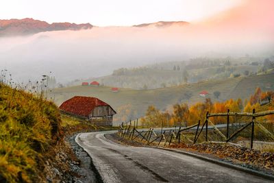 Countryside road by foggy mountain against sky at sunrise during autumn