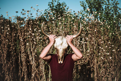 Full length of anonymous male in casual wear covering face with horn animal skull while standing in field