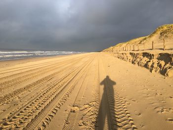 Tire tracks and shadow of person on sand at beach against sky