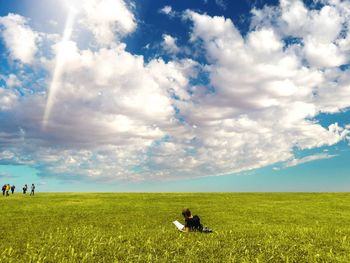 Man reading book on grassy field against cloudy sky