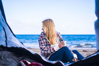 Woman sitting with coffee cup at beach against sky seen from tent