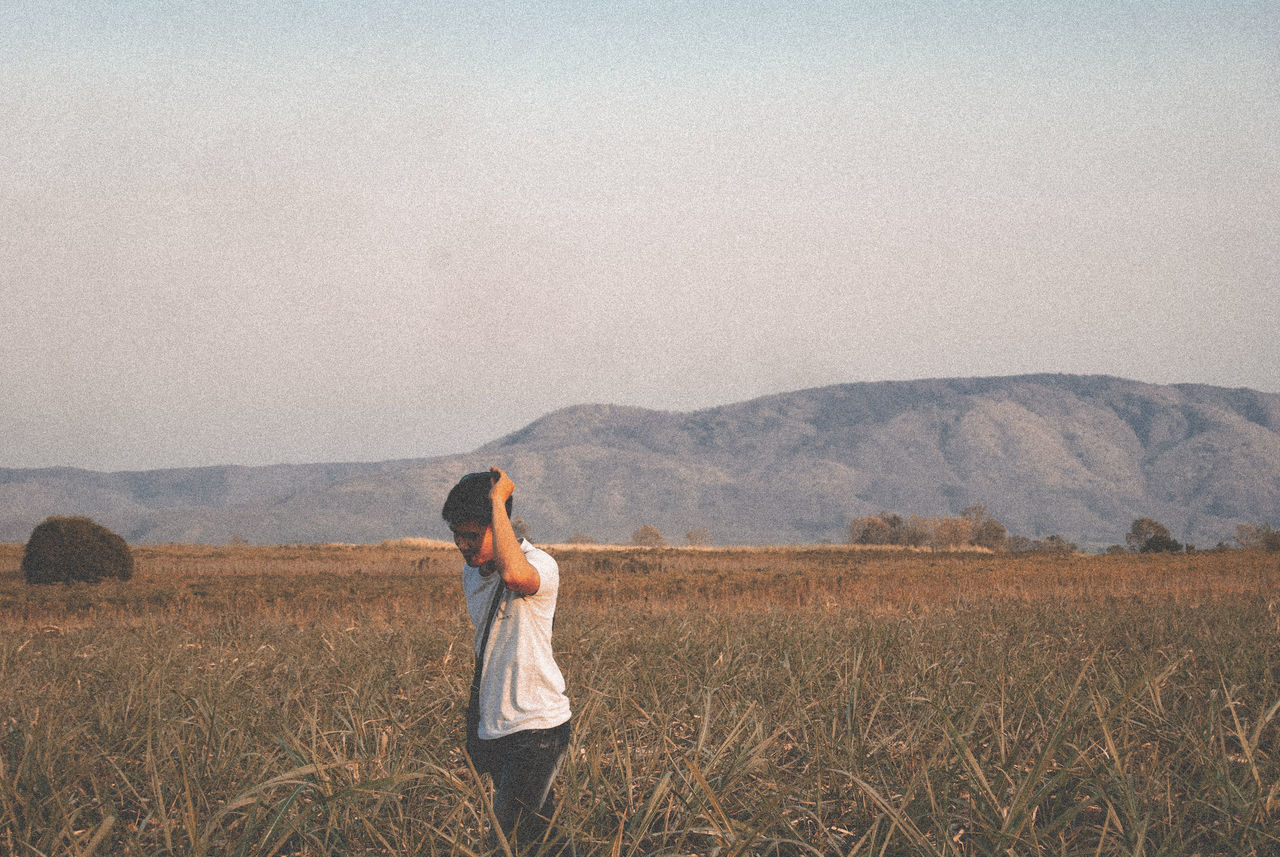 one person, field, land, landscape, standing, sky, environment, mountain, nature, scenics - nature, casual clothing, beauty in nature, leisure activity, young adult, non-urban scene, tranquility, day, real people, plant, outdoors, hairstyle