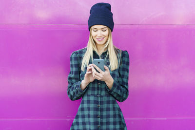 Young woman using phone while standing against pink wall