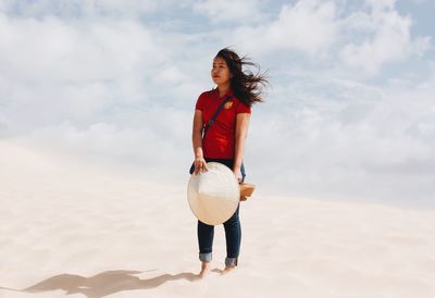 Woman holding asian style conical hat while standing in desert against sky during sunny day