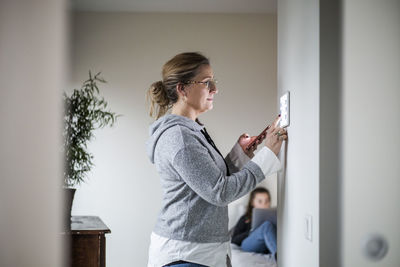 Mother holding smart phone while using digital tablet on wall with daughter in background at smart home