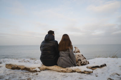 Young couple sitting with australian shepherd dog on snow covered log at beach