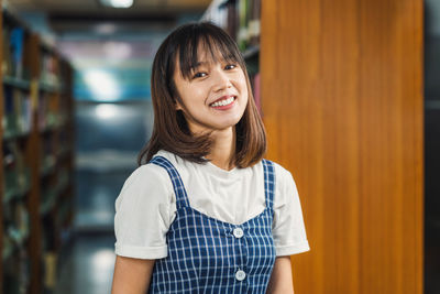 Portrait of smiling young woman standing against shelf