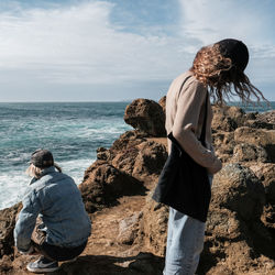 Rear view of boy sitting on rock by sea against sky