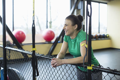 Female instructor smiling and looking away while leaning on chainlink fence in gym