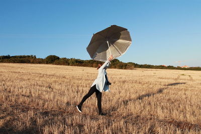 Woman with arms raised holding umbrella in field against clear sky during sunny day