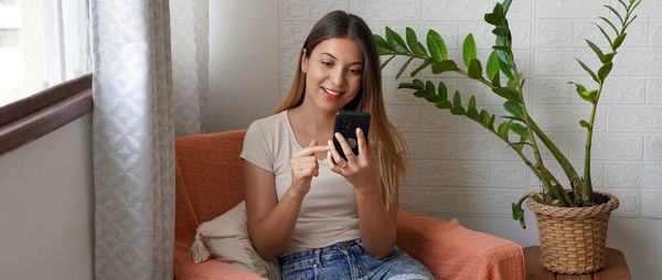 Panoramic banner of cute brazilian girl sitting on armchair using her smartphone doing e-commerce 