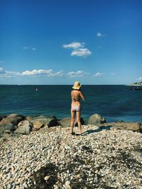 Rear view of young woman in bikini standing at beach against blue sky during sunny day