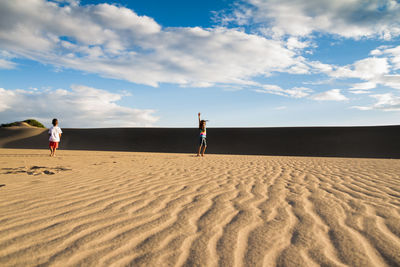 Siblings walking on sand dune at beach against sky