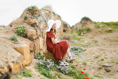 Cute caucasian kid girl in summer dress and white hat sits on stones with wild flowers. 