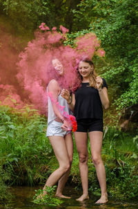 Full length of female friends with distress flares standing at lakeshore in forest