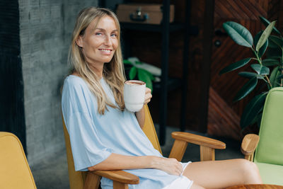 Happy smiling woman in a t-shirt and shorts sits in a coffee shop and drinks coffee.
