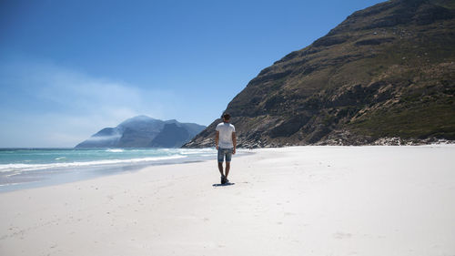 Rear view of man walking on sand at beach against blue sky during sunny day