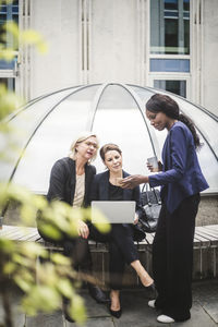 Full length of businesswoman showing smart phone to female coworkers in office yard