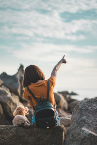 Rear view of woman sitting with dog on rocky shore against sky