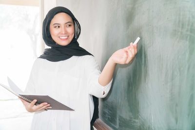 Smiling teacher wearing traditional clothing writing on blackboard while standing in classroom