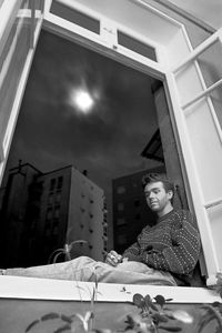 Low angle view of thoughtful young man smoking cigarette while sitting on window sill at home