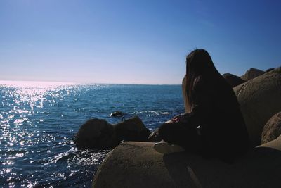 Woman sitting on rock looking at sea against clear sky