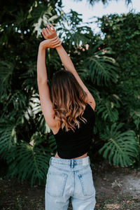 Back view of slender female in casual outfit standing with raised arms and stretching body in tropical garden with green monstera plants