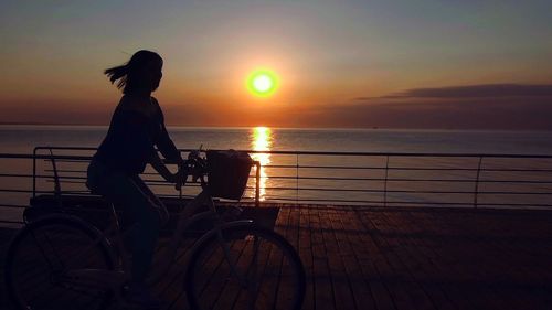 Silhouette woman riding bicycle on promenade against sky during sunset