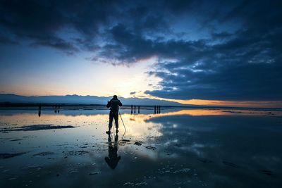 Rear view of photographer on wet shore against sky during sunset