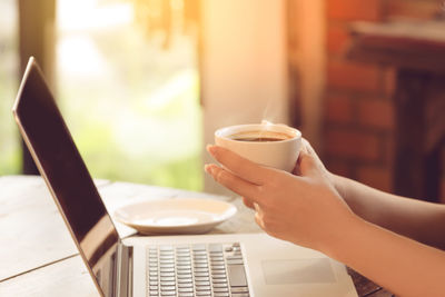 Cropped image of woman holding coffee cup while using laptop