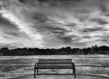 Empty bench on riverbank against cloudy sky