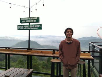 Portrait of young man standing on railing against sky